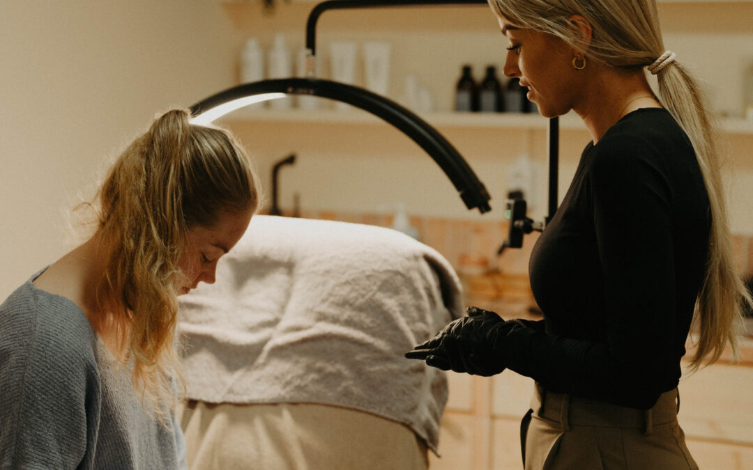 De afbeelding toont Ilse, een professionele PMU-artieste, terwijl ze de voorwaarden en adviezen rondom permanente make-up (PMU) uitlegt aan haar klant. Ilse staat of zit in een goed ingerichte salon, gekleed in professionele kledij, met een vriendelijke maar serieuze uitdrukking. Ze wijst op een document of tablet waarop de voorwaarden en adviezen staan vermeld. De klant, aandachtig luisterend, zit of staat tegenover Ilse, wat de interactieve en informatieve aard van het gesprek benadrukt. De salonomgeving is schoon en georganiseerd, met zichtbare PMU-benodigdheden op de achtergrond. Deze afbeelding legt de nadruk op het belang van goede voorlichting en communicatie tussen PMU-specialisten en hun klanten, wat essentieel is voor een veilige en tevredenstellende PMU-ervaring.