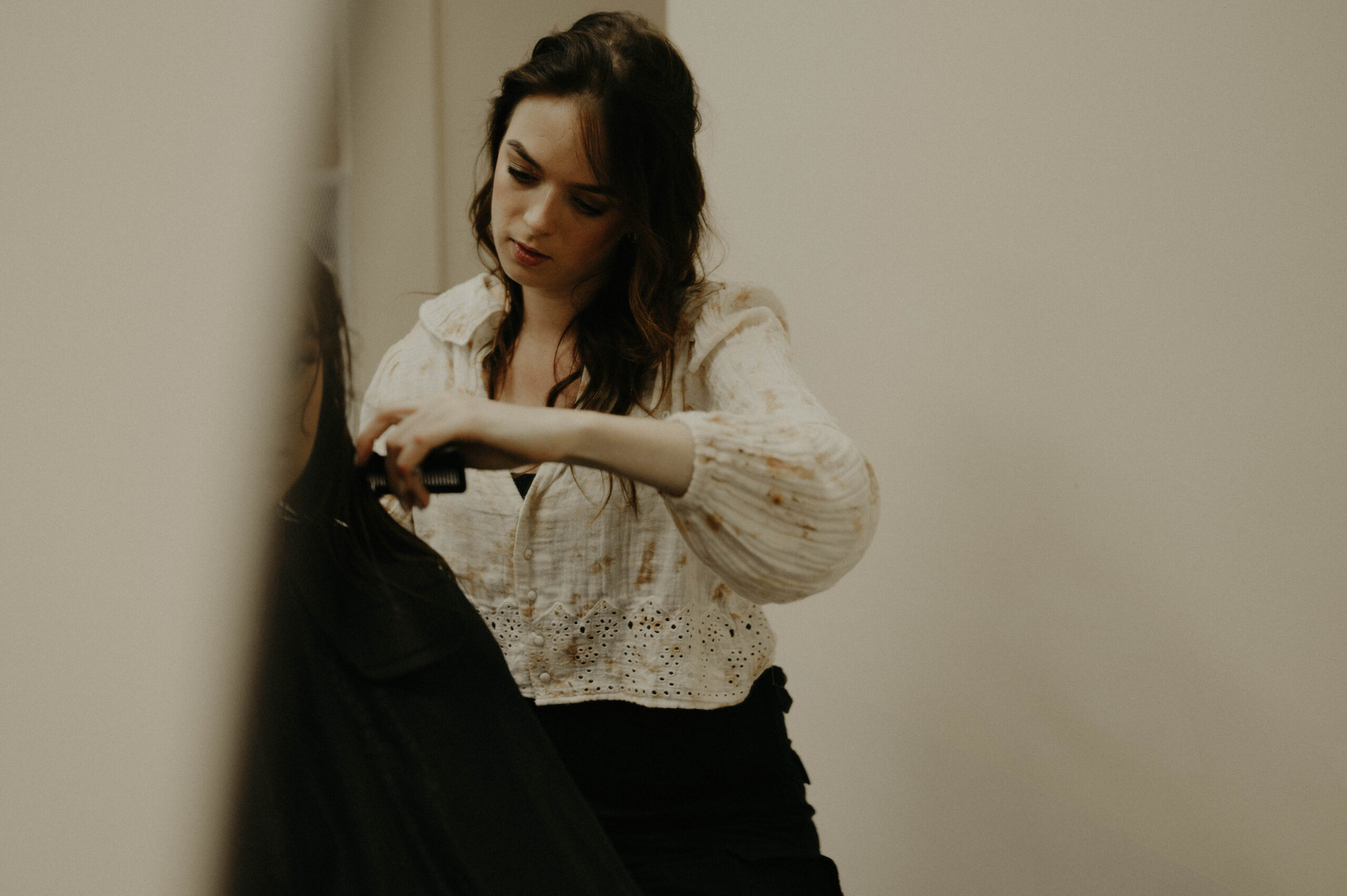Demi Demi, haarstylist bij WINO beauty lab, geconcentreerd aan het werk terwijl ze het haar van een klant knipt, gekleed in een lichte blouse met borduursels in een sfeervolle, warme salonomgeving.