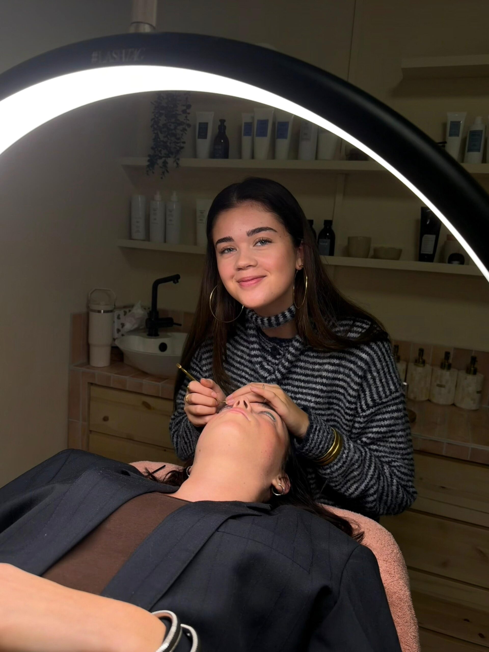 Foto van Demi, onze getalenteerde kapster bij WINO beauty lab, met een hartelijke glimlach terwijl ze ontspannen zit in de salon. Demi is stijlvol gekleed in een crèmekleurige, luchtige blouse en zwarte broek, uitkijkend naar de volgende klant die ze gaat helpen transformeren met haar expertise in hairstyling.