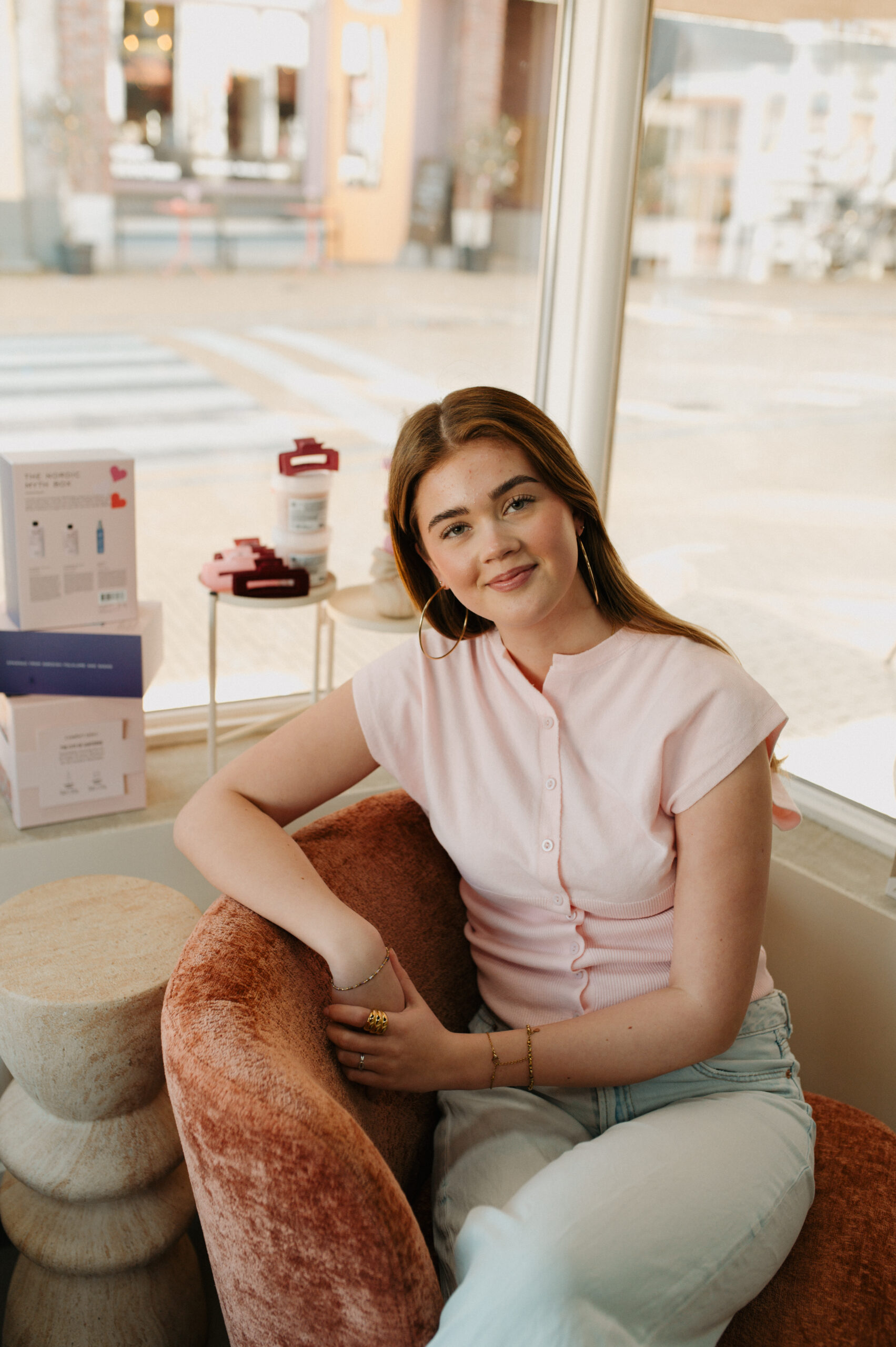 Foto van Demi, onze getalenteerde kapster bij WINO beauty lab, met een hartelijke glimlach terwijl ze ontspannen zit in de salon. Demi is stijlvol gekleed in een crèmekleurige, luchtige blouse en zwarte broek, uitkijkend naar de volgende klant die ze gaat helpen transformeren met haar expertise in hairstyling.