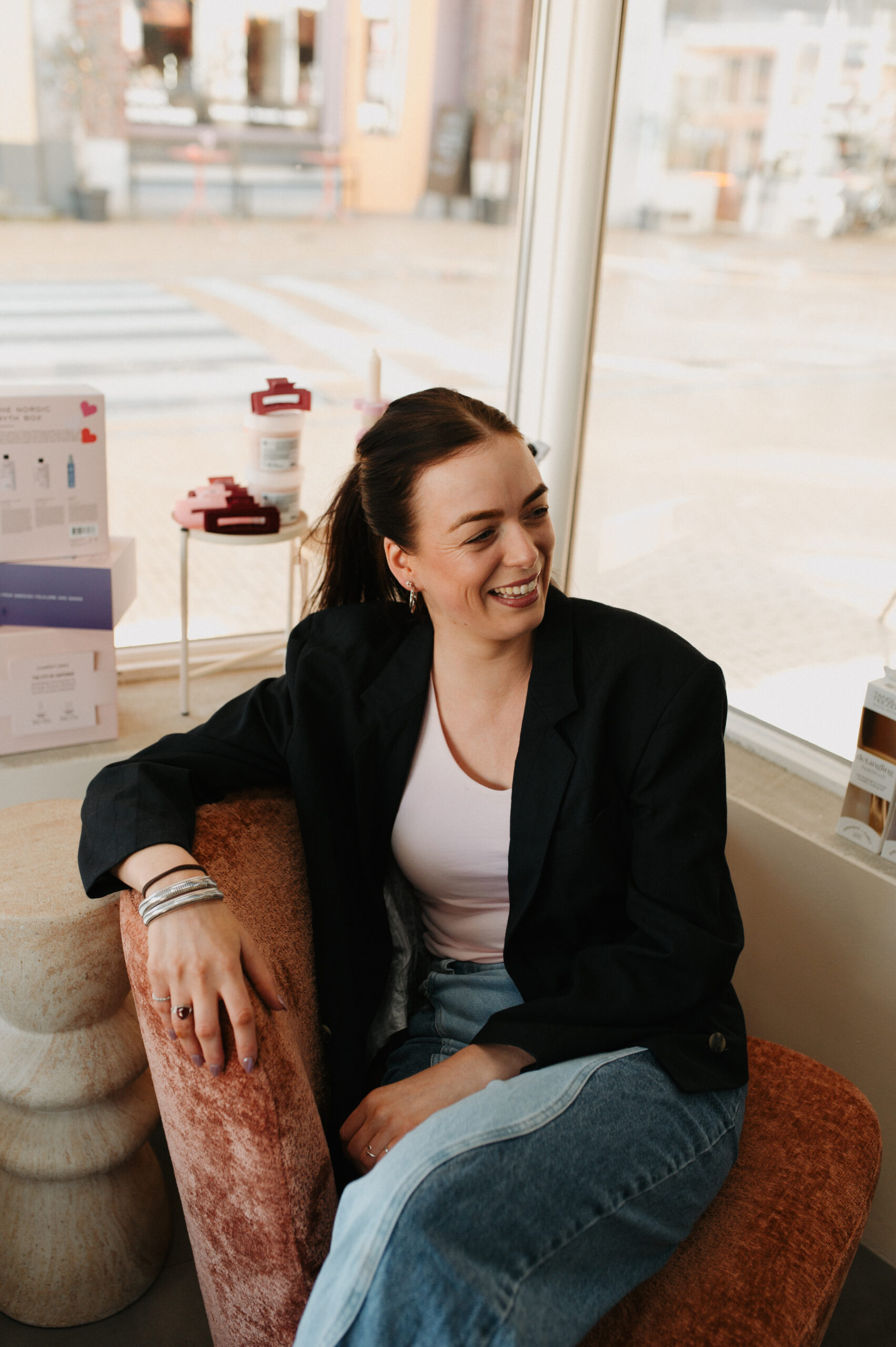 Foto van Demi, onze getalenteerde kapster bij WINO beauty lab, met een hartelijke glimlach terwijl ze ontspannen zit in de salon. Demi is stijlvol gekleed in een crèmekleurige, luchtige blouse en zwarte broek, uitkijkend naar de volgende klant die ze gaat helpen transformeren met haar expertise in hairstyling.